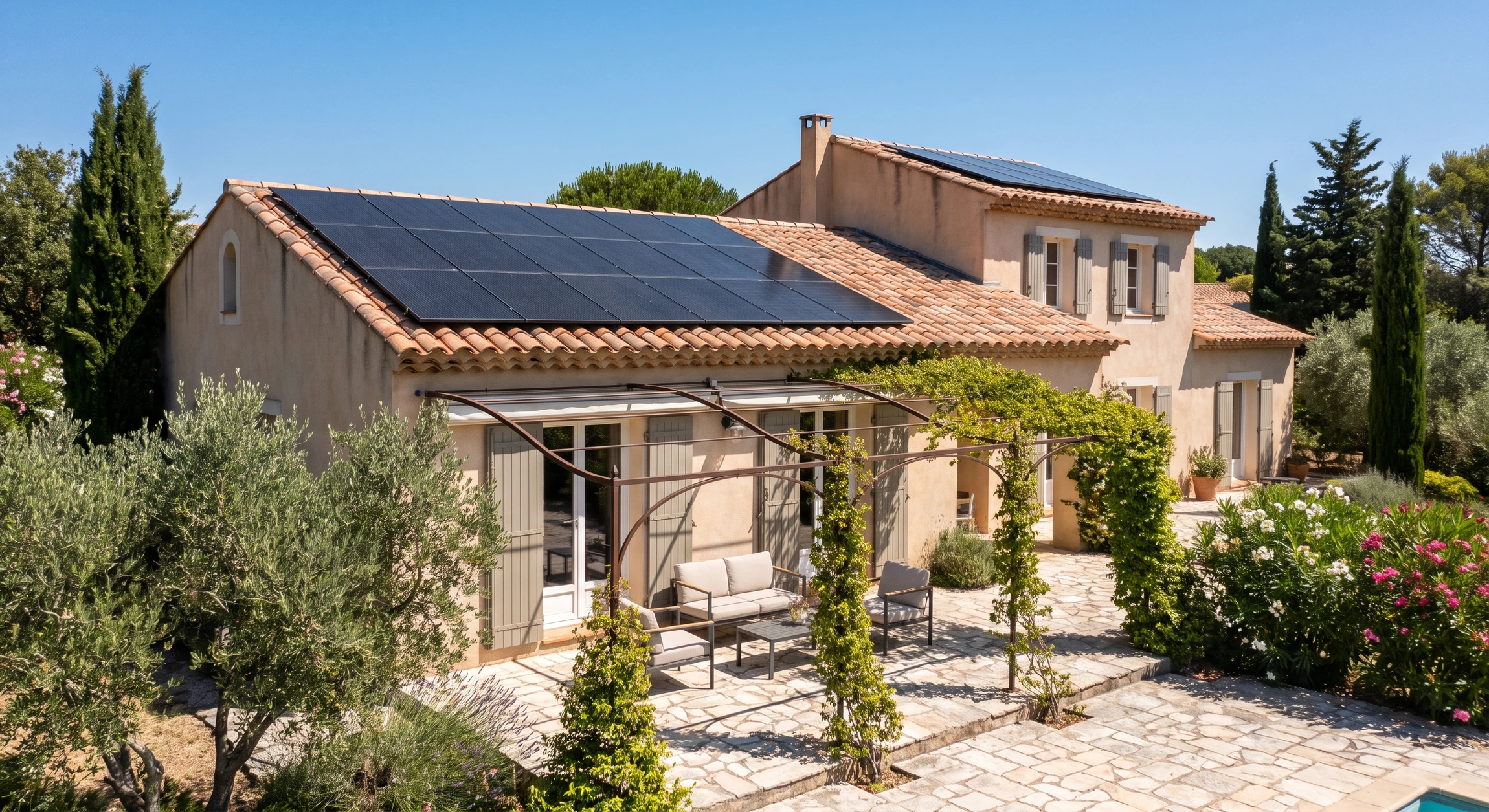 Maison résidentielle méditerranéenne avec toiture en tuiles canal terre cuite, équipée de panneaux solaires photovoltaïques monocristallins noirs en surimposition, terrasse dallée avec pergola en fer forgé noir, mobilier extérieur discret, environnement ensoleillé.